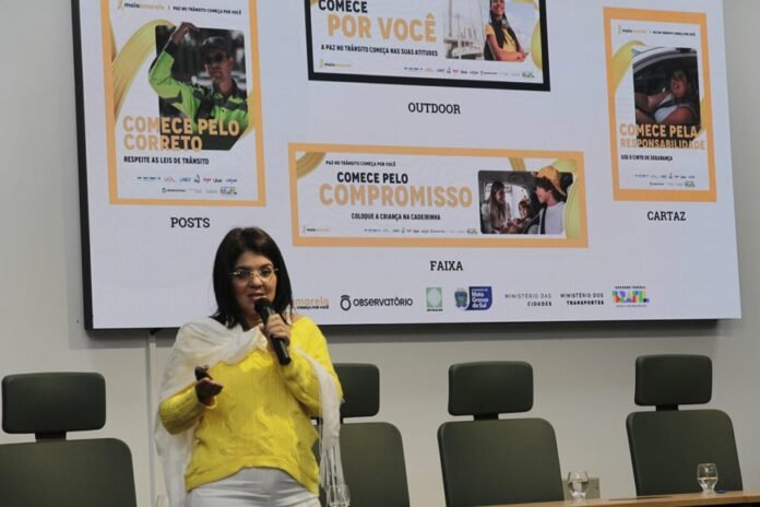 A woman in a yellow top speaks into a microphone while presenting, standing in front of a large screen showing posters and banners for a campaign or event.