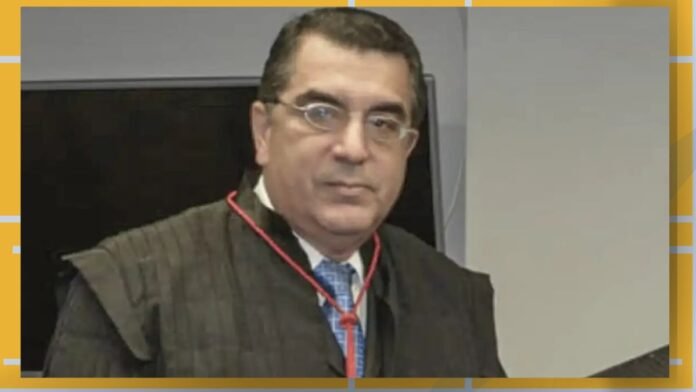 Man wearing a dark academic gown and blue tie, looking at the camera in a formal setting