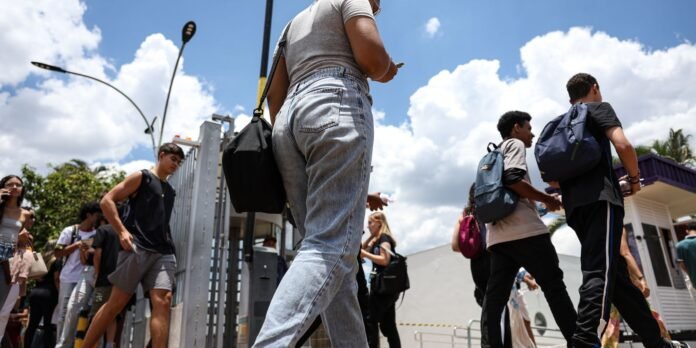 Group of students and pedestrians walking outside on a sunny day, seen from a low angle near a metal fence and buildings behind them.