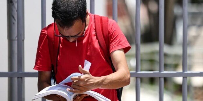 Man in a red t-shirt with a backpack reads an open book outdoors beside a metal railing.