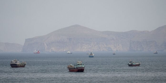 Several cargo ships afloat on calm sea with pale, rocky cliffs and mountains in the hazy background.