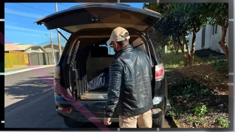 Man wearing a black leather jacket and beige cap stands at the open tailgate of an SUV, loading a large wrapped item into the cargo area on a sunny street.