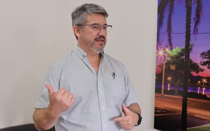 Man with gray hair and glasses speaks, gesturing with thumbs up and other hand, wearing a light button-down shirt; wall and a sunset palm-tree mural in background.