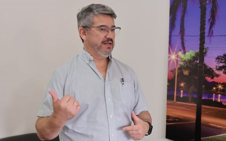 Man with gray hair and glasses speaks, gesturing with thumbs up and other hand, wearing a light button-down shirt; wall and a sunset palm-tree mural in background.