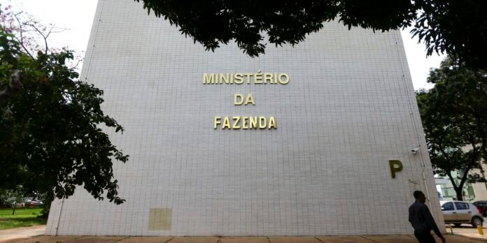 Facade of Ministério da Fazenda building with white tiles and gold letters; a man walks by on the right with trees framing.