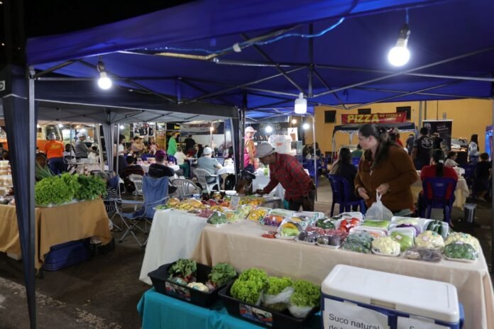 Night market scene under blue canopies with vendors selling fresh vegetables and prepared foods; people browse and dine at tables under strings of lights.