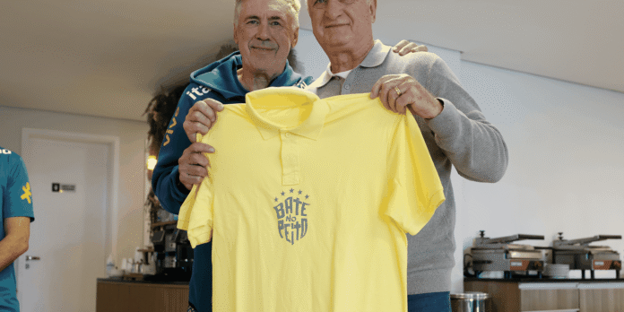 Two older men stand side by side, smiling, while holding up a bright yellow polo shirt with a circular star logo. in a room with service equipment in the background.