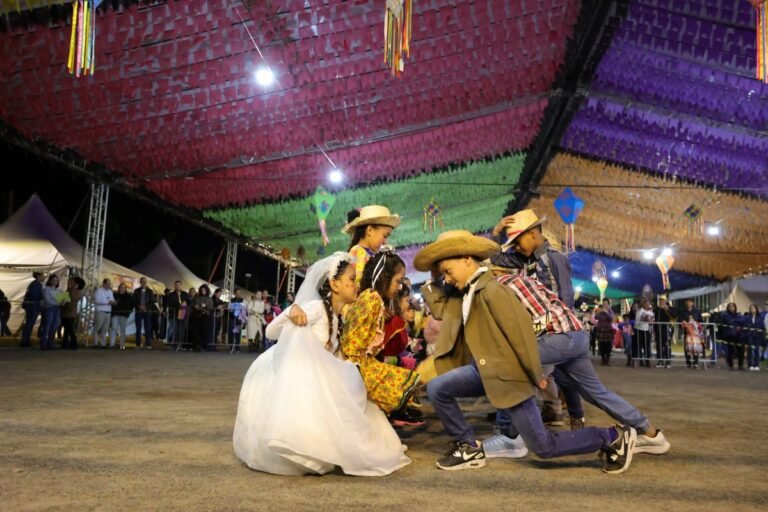 Children in colorful traditional outfits and cowboy hats dance in a festival square as adults watch from behind barriers under bright, multicolored banners.