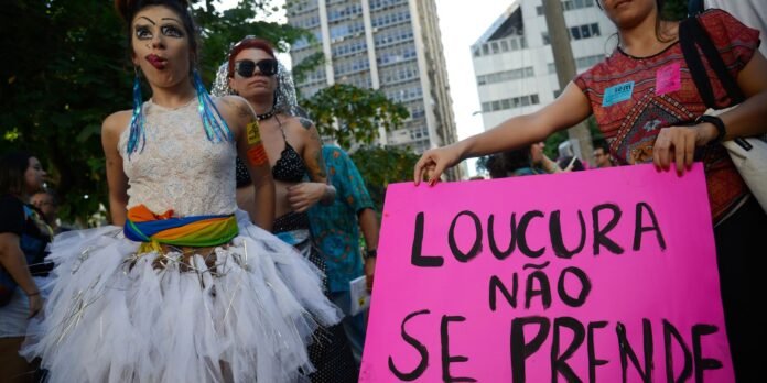 Drag performer in a white lace dress with a rainbow sash at a street protest, next to a bright pink sign reading 'LOUCURA NÃO SE PRENDE'.
