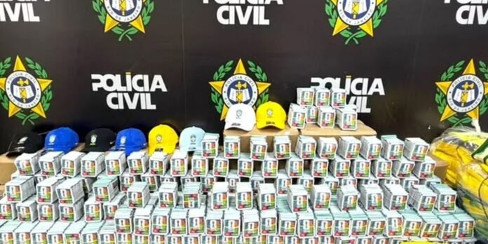 Row of white boxed items with colorful labels arranged on a table in front of a Polícia Civil backdrop, with blue, black, white, and yellow caps on top and yellow bags to the right.