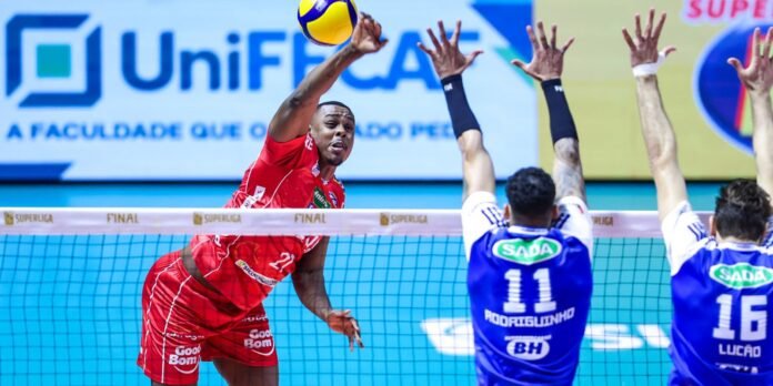 Volleyball player in a red uniform spikes the ball over the net as blue-team blockers reach up to block it.