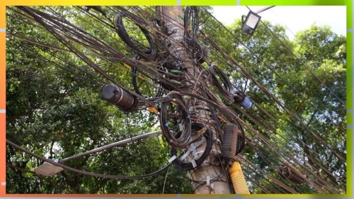 A utility pole overloaded with a tangle of electrical wires and cables, with a streetlight and green trees in the background.