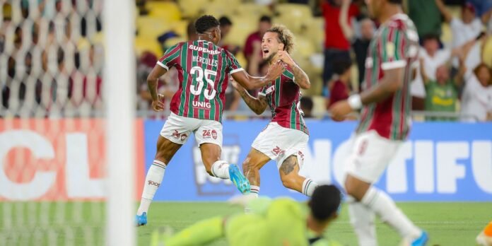 Two Fluminense players in green, maroon and white celebrate a goal near the net as the goalkeeper lies on the field in the foreground.