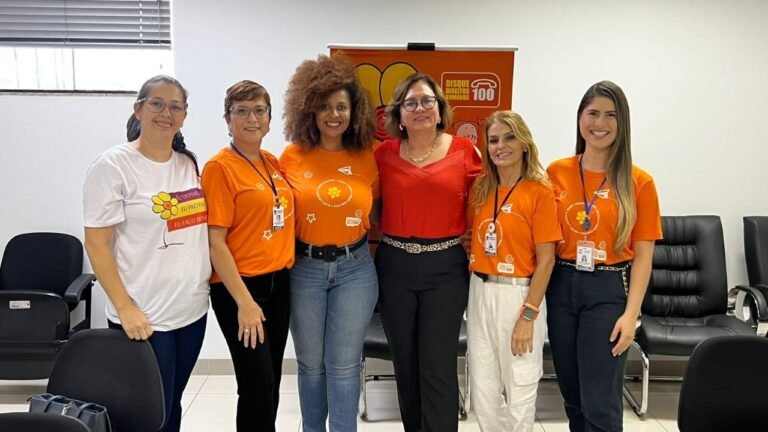 Six women posing for a group photo in an office, five wearing orange shirts with name badges and a banner in the background.