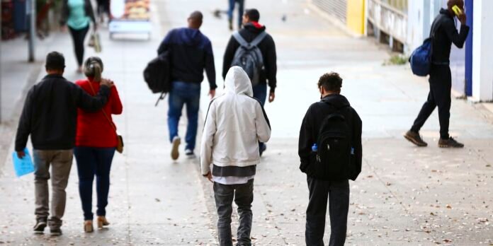People walk down a city sidewalk from behind; two individuals in foreground wear hoodies and backpacks, while others stroll nearby