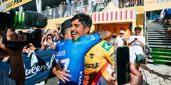 Two cyclists embrace in a victory hug on a sunny beach-boardwalk as fans cheer and photographers capture the moment.