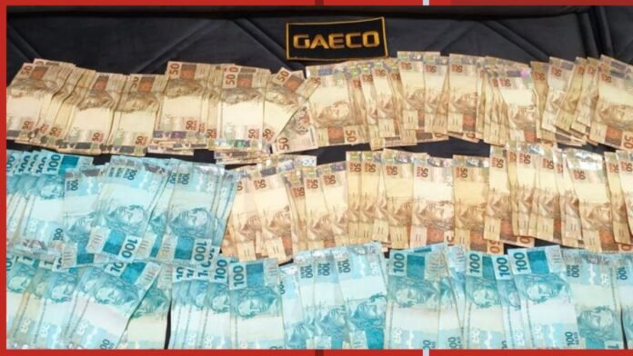 Stacks of Brazilian real banknotes in beige and blue tones arranged on a dark surface with a GAEC0 label visible on top.