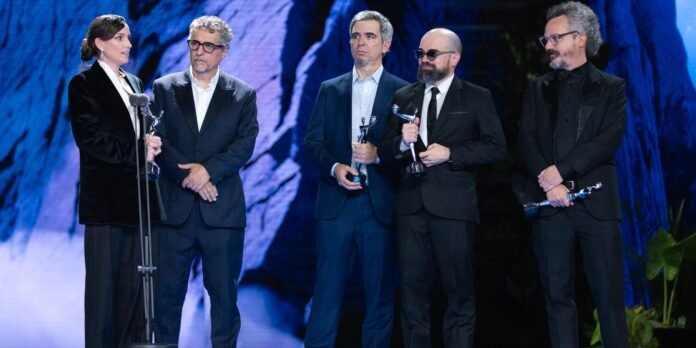 Five people in dark suits stand on a stage, each holding a trophy during an awards ceremony with a blue-lit backdrop and a microphone nearby.
