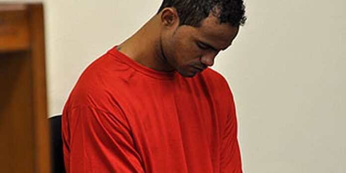 man in an orange jumpsuit looking down, in a detention or courtroom setting