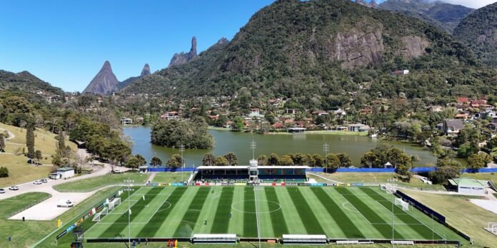 Aerial view of a soccer stadium beside a lake, with a striped green pitch and surrounding houses and forested mountains in the background.