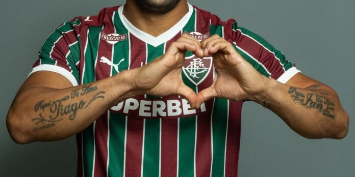 Soccer player in a Fluminense jersey forms a heart shape with hands over the chest; visible forearm tattoos.