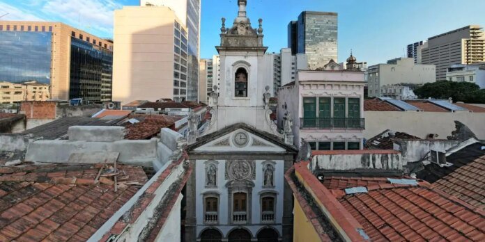 Historic church with ornate facade rising between weathered red-tile roofs and a backdrop of modern skyscrapers behind it.
