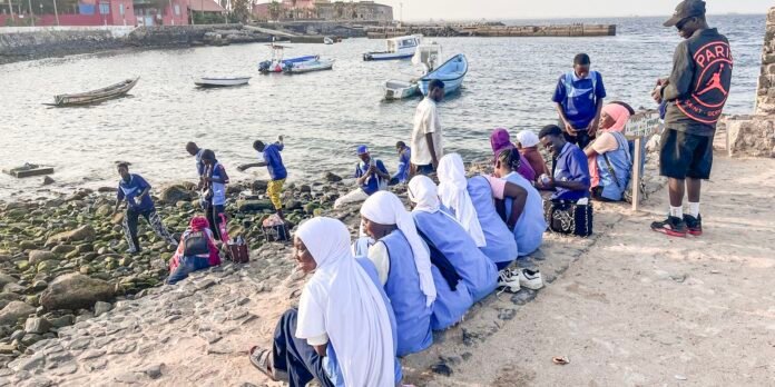 Group of students in blue uniforms and white headscarves sit along a rocky seawall by a harbor, while others explore the shoreline and boats sit in the water behind them.