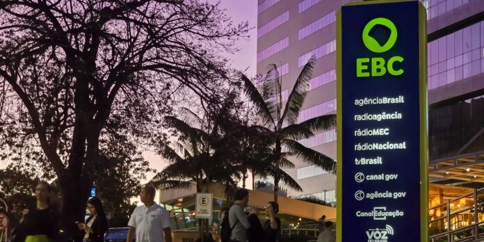 Vertical EBC sign listing Brazilian media agencies beside a glass-front building at dusk, with palm trees and pedestrians in foreground.