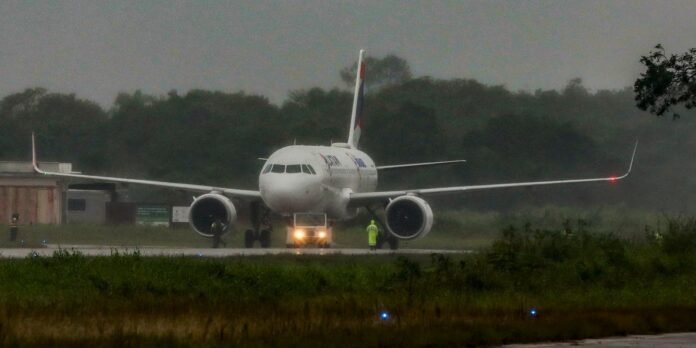Front view of a large white commercial airliner on a wet runway with a pushback tug in front and ground crew in high‑visibility jackets nearby, during overcast weather.