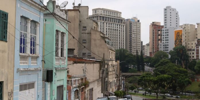 Colorful pastel row houses line a sloped street with modern high-rise buildings in the background and a mural on an orange building to the right.
