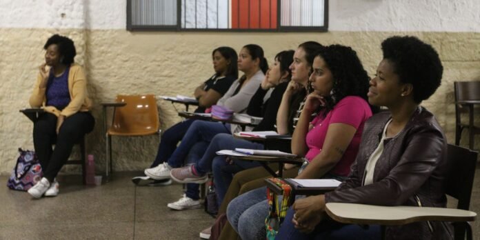 A row of adult students seated with notebooks in a classroom, listening to a lecturer on the left wearing a yellow cardigan.