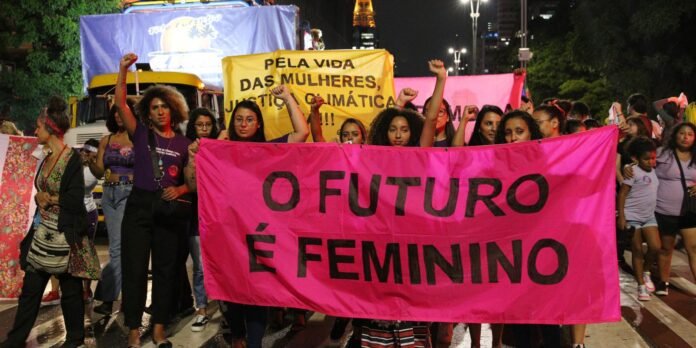 Group of diverse women marching at night holding a bright pink banner that reads 'O FUTURO É FEMININO'.