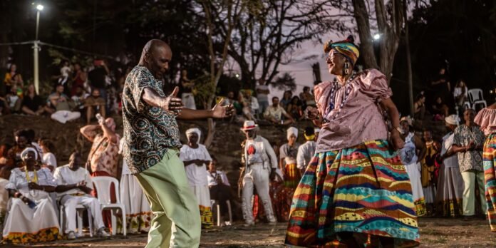 Dancing couple at an outdoor evening celebration; man in a patterned shirt and light pants, woman in a bright multicolored skirt and pink top, crowd in white attire watching in the background.