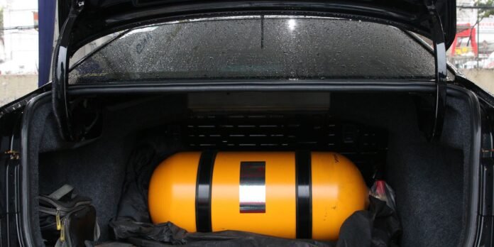 Orange cylindrical air tank strapped in the open trunk of a car, surrounded by bags and a black cloth on a rainy day.