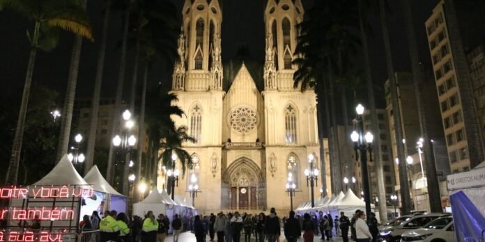 Night view of a illuminated Gothic cathedral with white vendor tents and a crowd at a night festival along a plaza.