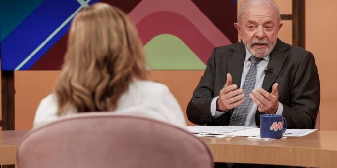 TV interview scene with an older man in a dark suit speaking to a person seen from behind, colorful geometric backdrop behind them.