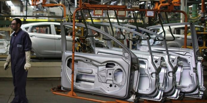 Factory assembly line with silver car door panels mounted on an orange rack, worker in coveralls nearby