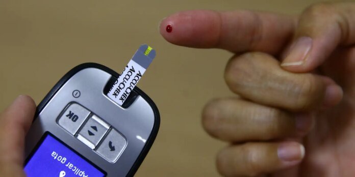 Close-up of a finger with a small blood drop beside a glucometer test strip being inserted into the device for a blood glucose test.