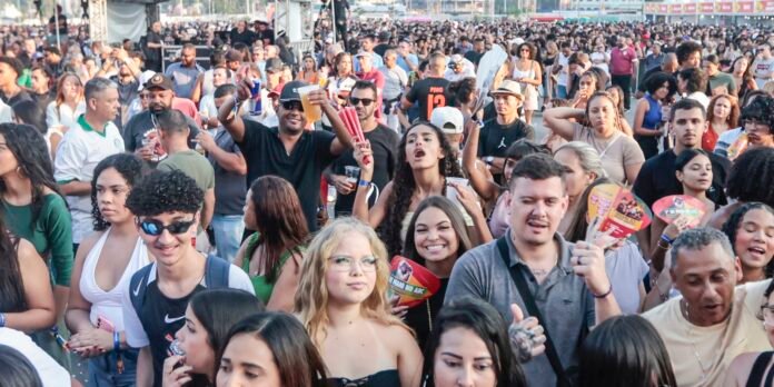 Large, diverse crowd at an outdoor concert or festival, people cheering and holding drinks and fans in a sunlit open area, with a stage in the background.