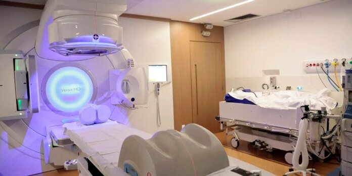 Hospital radiotherapy suite with a Versa HD linear accelerator and treatment couch prepared for a patient. The room includes monitoring screens and medical tubing.
