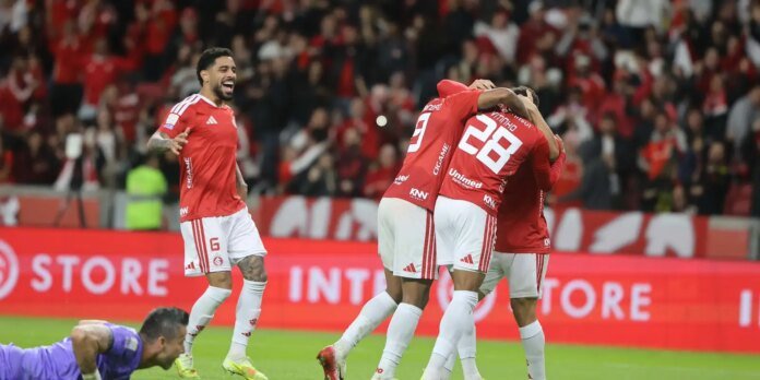 Soccer players in red uniforms celebrate a goal on the field with a cheering crowd in the stands behind them.