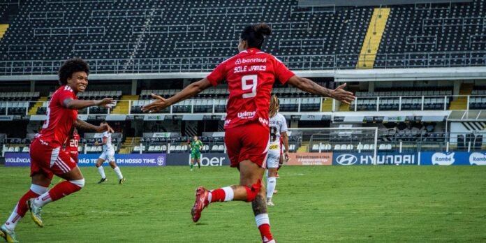 Soccer players in red jerseys celebrate a goal on a green field, central player #9 with arms outstretched as teammates approach. Empty stadium seats in background.
