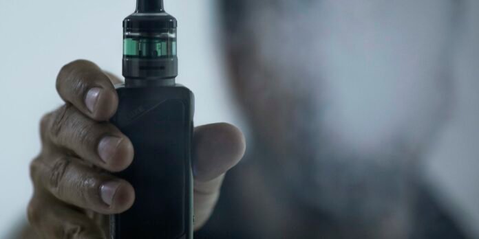Close-up of a hand gripping a black vape mod with a green-t liquid tank at the top, held ready to vape