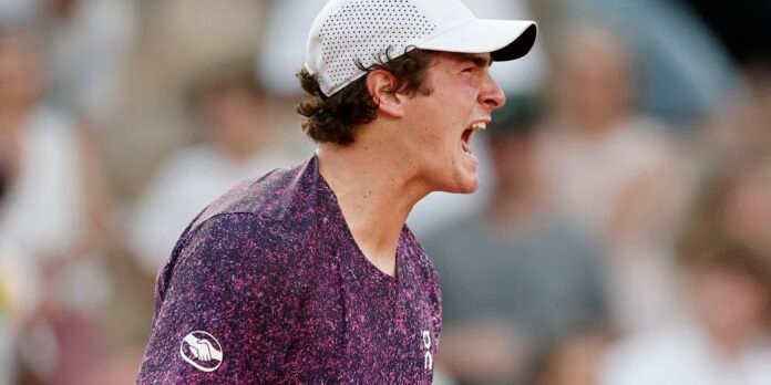 Male tennis player shouting on court, wearing a white cap and purple speckled shirt with a crowd in the background