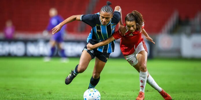 Two female soccer players chase the ball during a match; one wears a blue‑and‑black striped kit, the other red kit.