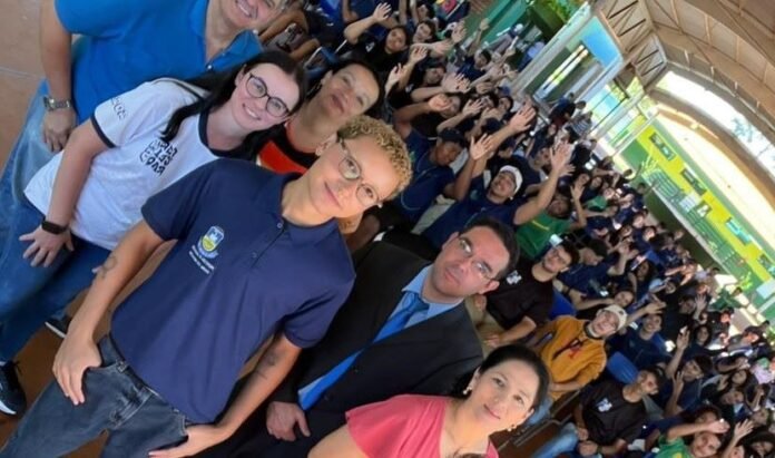 Large group of people posing for a selfie-style photo in an indoor venue, many smiling and waving at the camera.