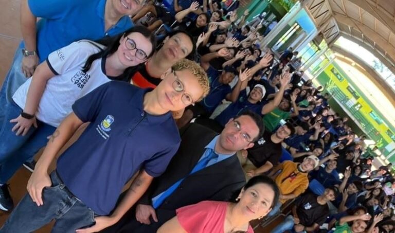 Large group of people posing for a selfie-style photo in an indoor venue, many smiling and waving at the camera.