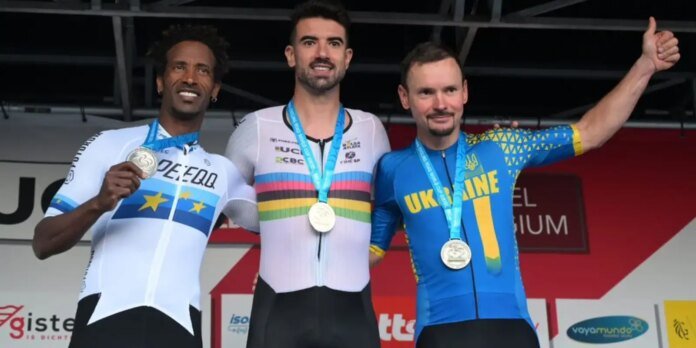Three male cyclists stand on a podium with medals around their necks: left in white/blue, center in a rainbow-striped jersey, right in blue and yellow Ukraine kit, giving a thumbs-up.