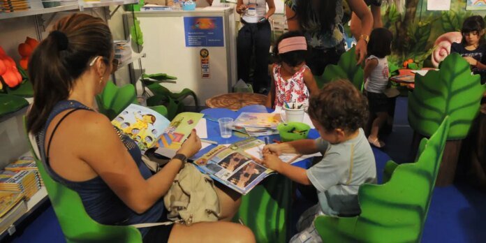 Adult and child seated at a blue table reading and drawing from colorful magazines in a leaf-themed kids' activity area.