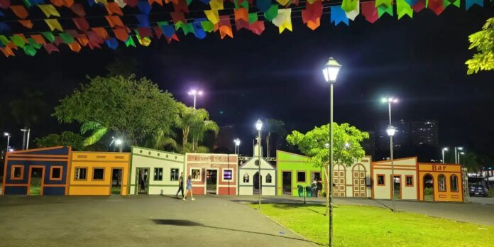 Nighttime view of a colorful row of small decorative buildings forming a plaza with string festival banners overhead and street lamps